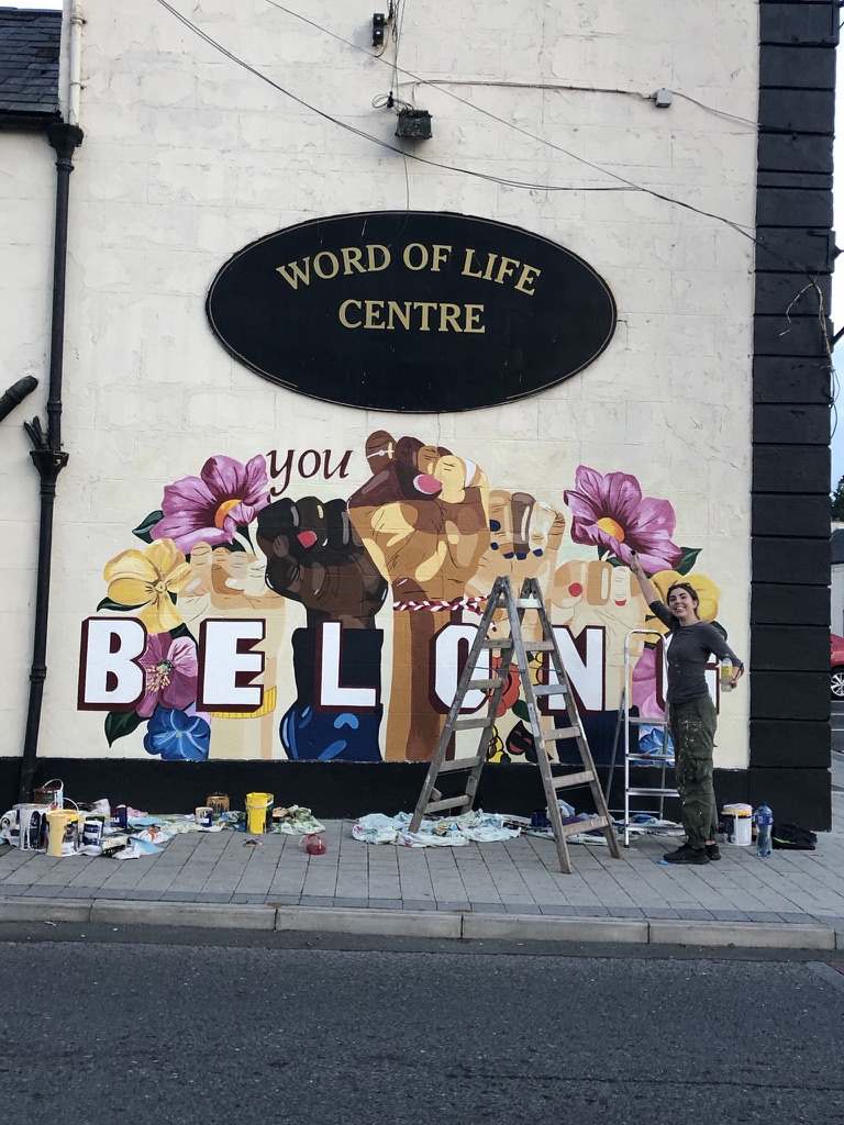 Aisling Corcoran painting a large outdoor mural with raised fists and flowers that reads You Belong